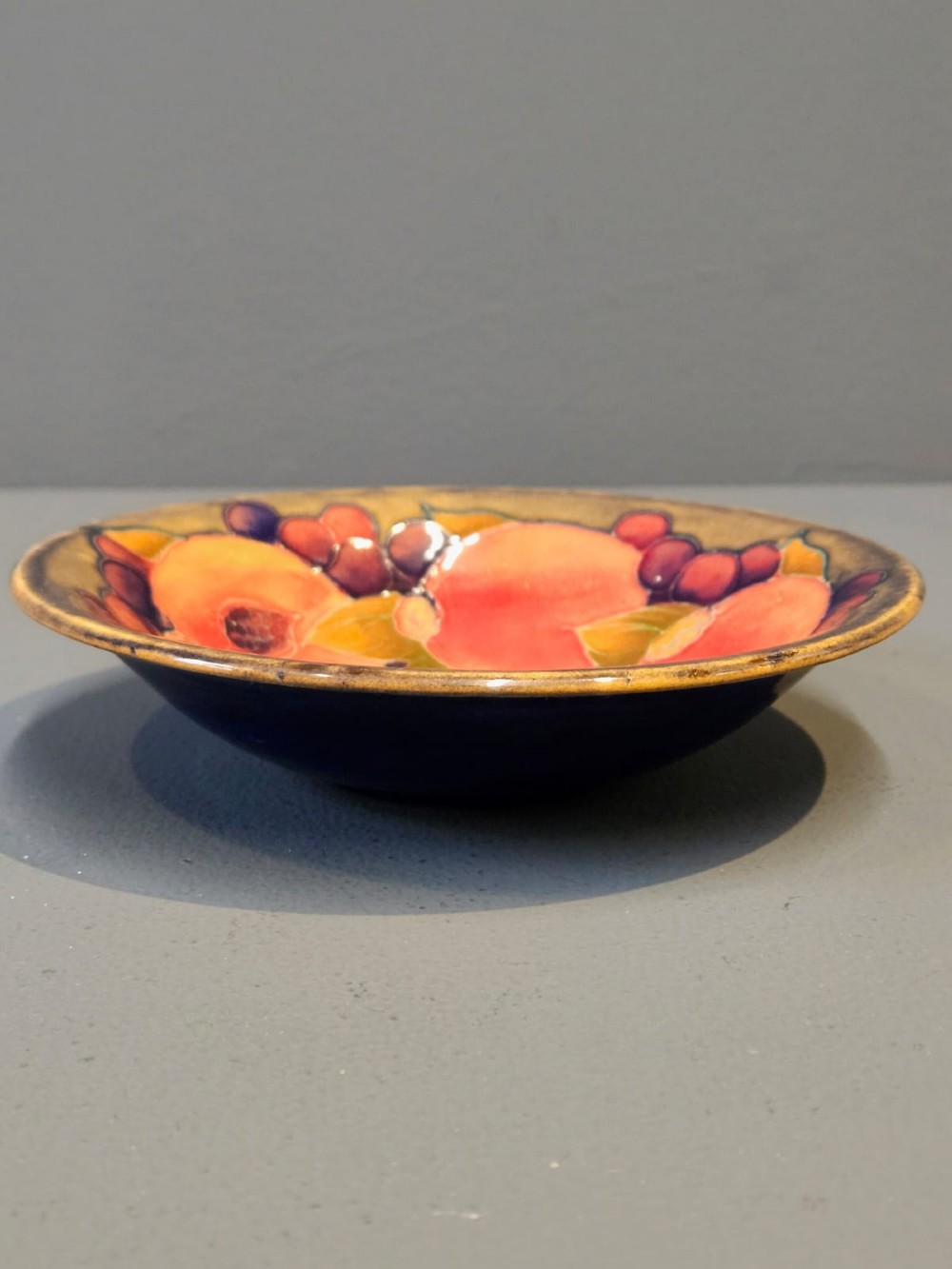 A circular shallow earthenware bowl decorated with hand-painted tubelined pomegranate fruits and dark berries over an ochre-tinted ground. View II