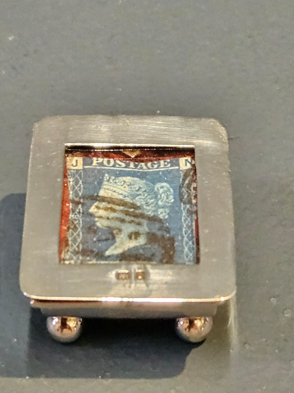 An Edwardian sterling silver stamp box featuring four ball feet and a glass inset lid displaying an antique postage stamp.