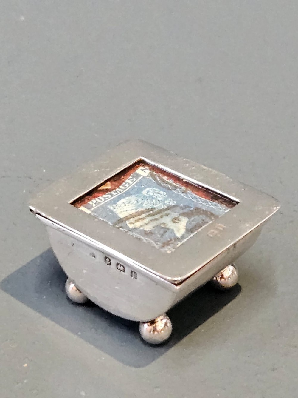An Edwardian sterling silver stamp box featuring four ball feet and a glass inset lid displaying an antique postage stamp. View II