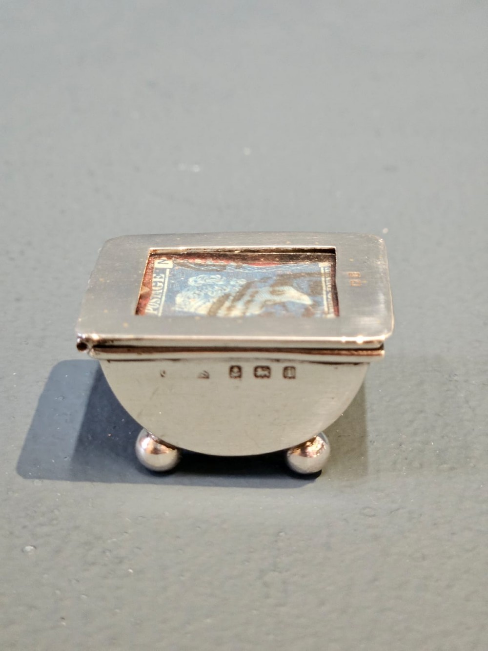 An Edwardian sterling silver stamp box featuring four ball feet and a glass inset lid displaying an antique postage stamp. View III