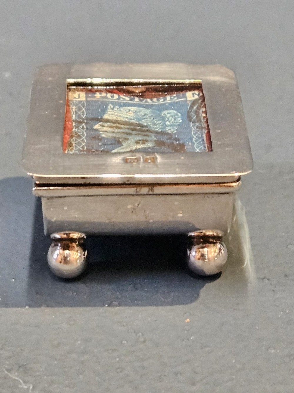 An Edwardian sterling silver stamp box featuring four ball feet and a glass inset lid displaying an antique postage stamp. View IV