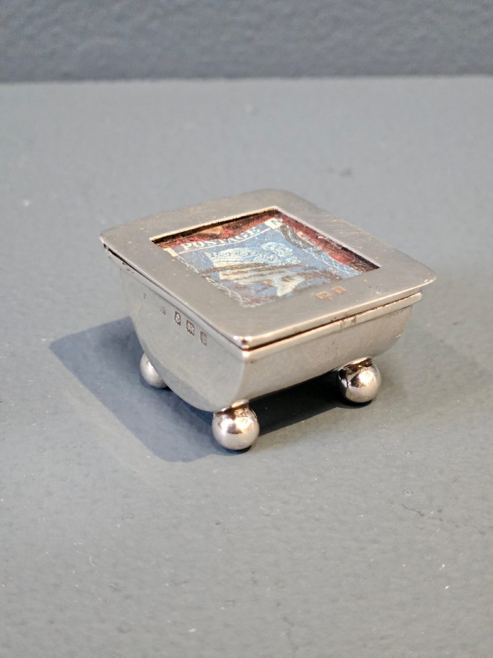 An Edwardian sterling silver stamp box featuring four ball feet and a glass inset lid displaying an antique postage stamp. View V