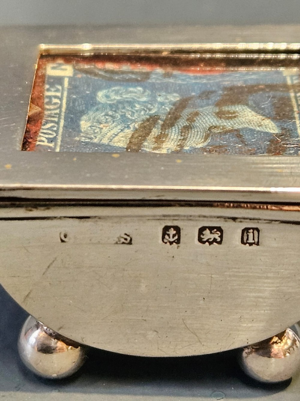 An Edwardian sterling silver stamp box featuring four ball feet and a glass inset lid displaying an antique postage stamp. View VI