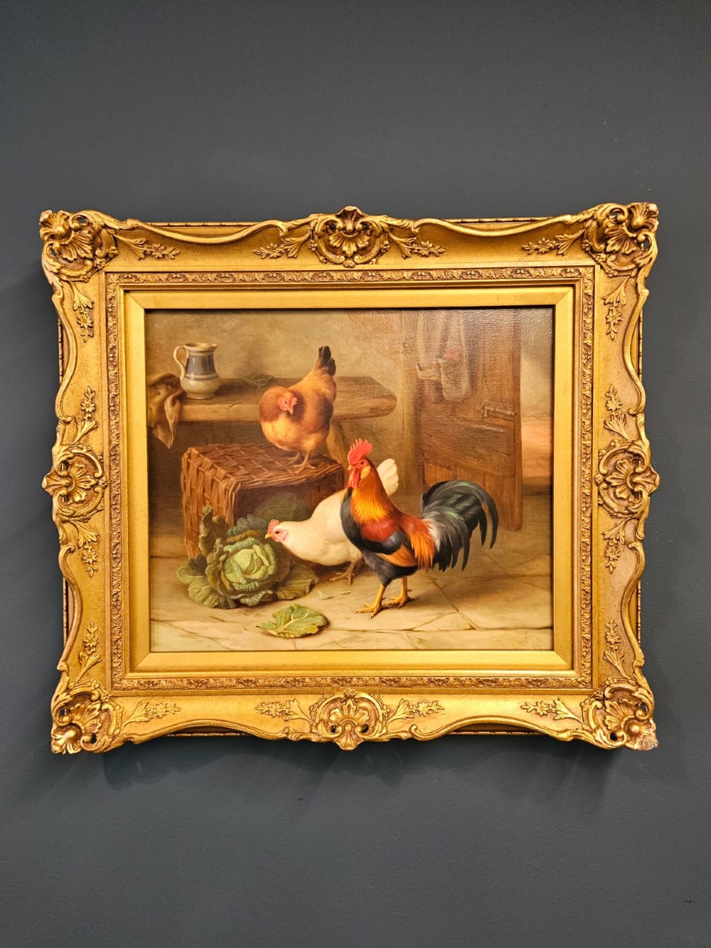 An oil painting on canvas depicting a cockerel and two hens inside a rustic barn beside a wicker basket and cabbage, enclosed in a heavily carved gold frame.