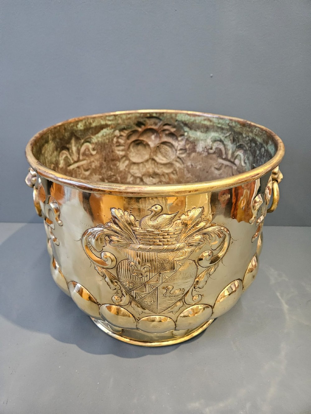A large polished brass cylindrical planter with an embossed heraldic crest on the front, godrooned base pattern, and side ring handles.