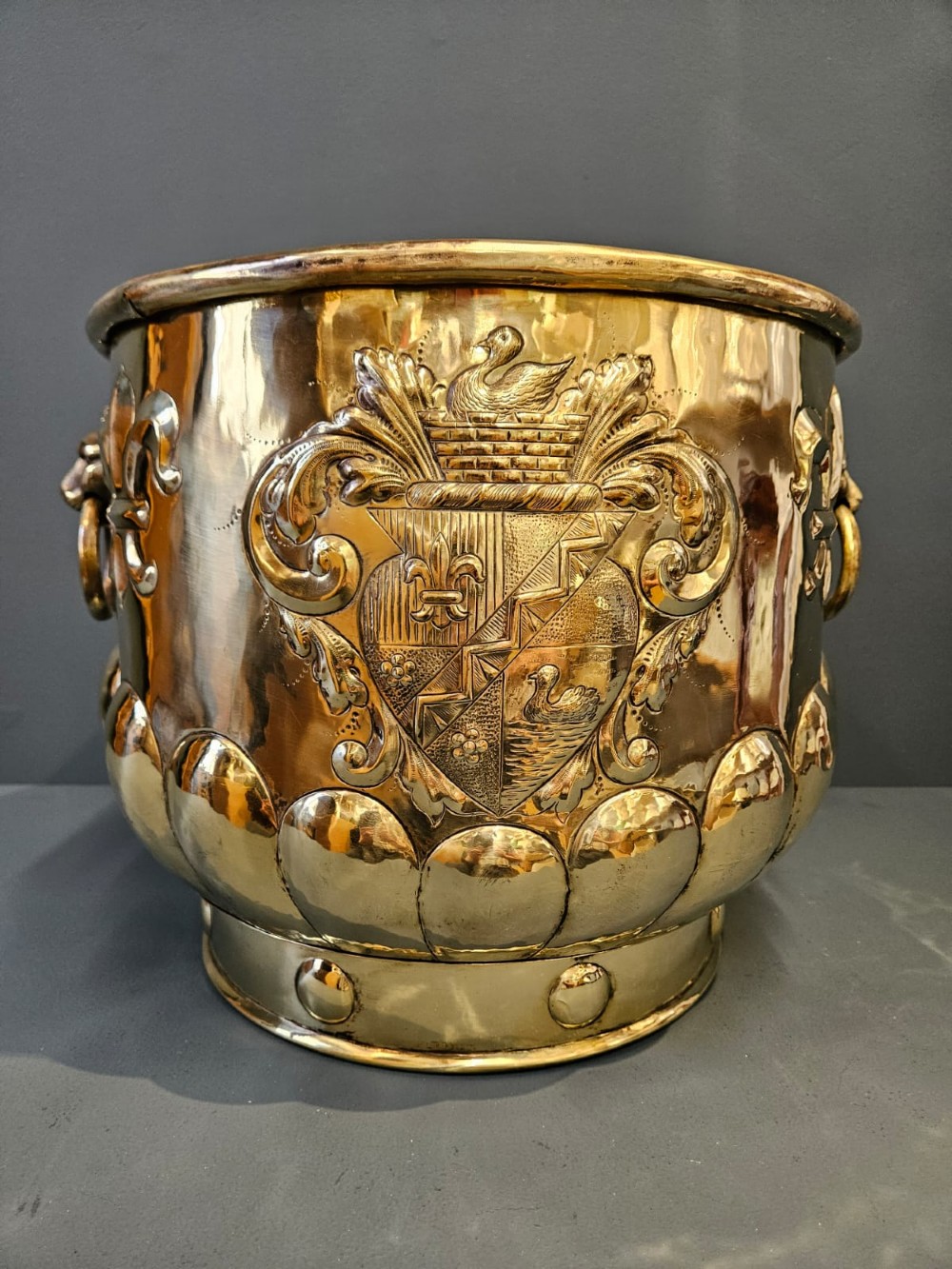A large polished brass cylindrical planter with an embossed heraldic crest on the front, godrooned base pattern, and side ring handles. View III