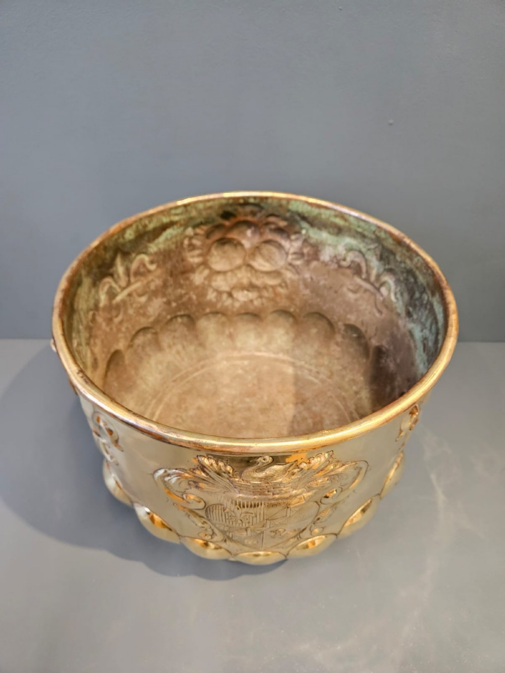 A large polished brass cylindrical planter with an embossed heraldic crest on the front, godrooned base pattern, and side ring handles. View IV