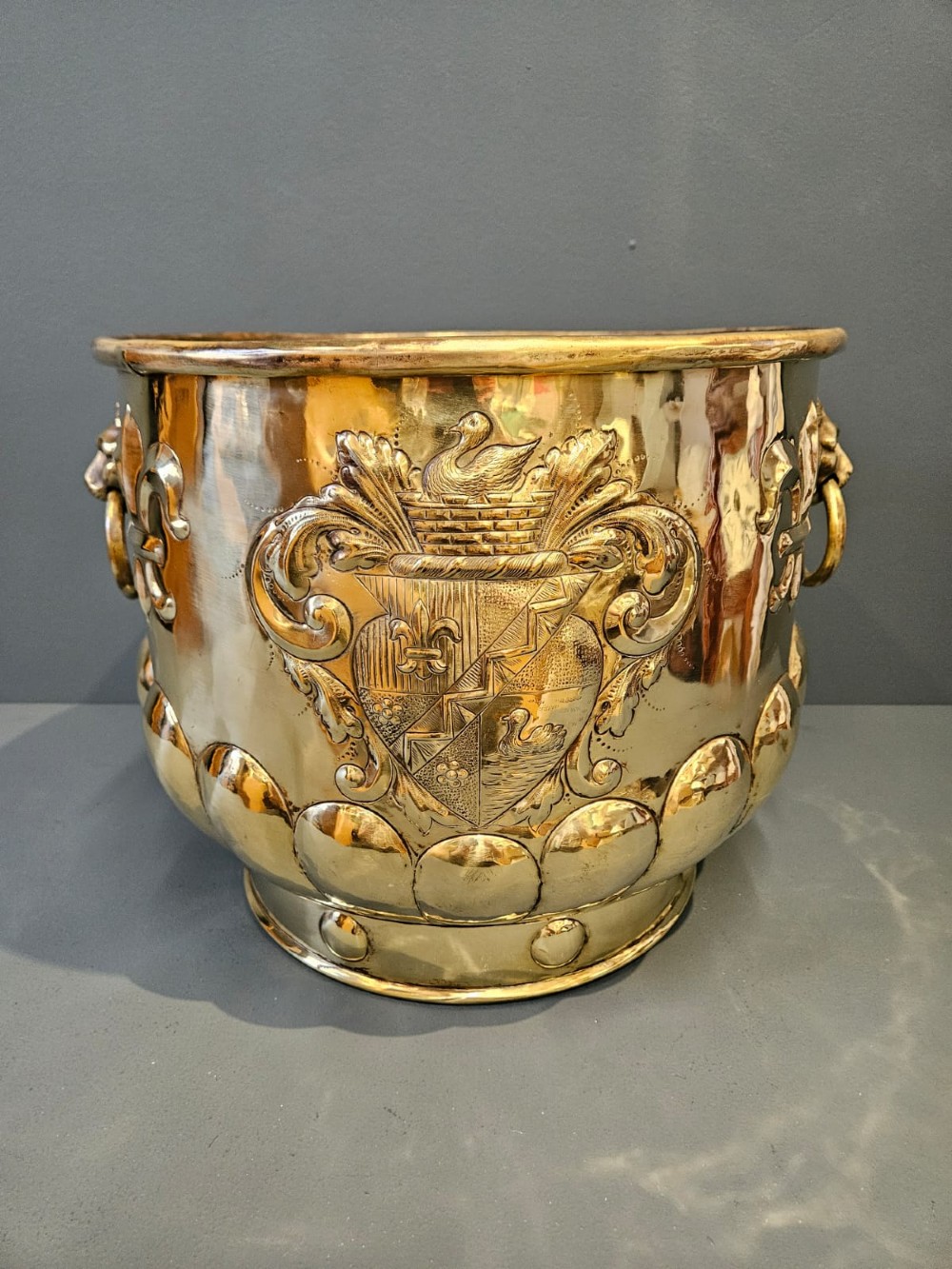 A large polished brass cylindrical planter with an embossed heraldic crest on the front, godrooned base pattern, and side ring handles. View VI
