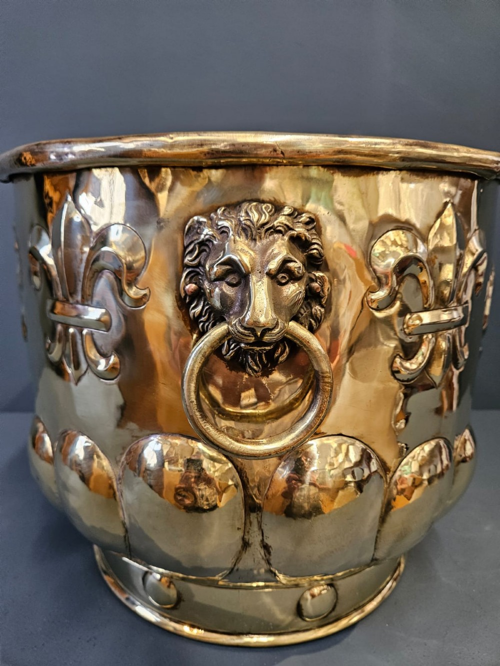 A large polished brass cylindrical planter with an embossed heraldic crest on the front, godrooned base pattern, and side ring handles. View VII