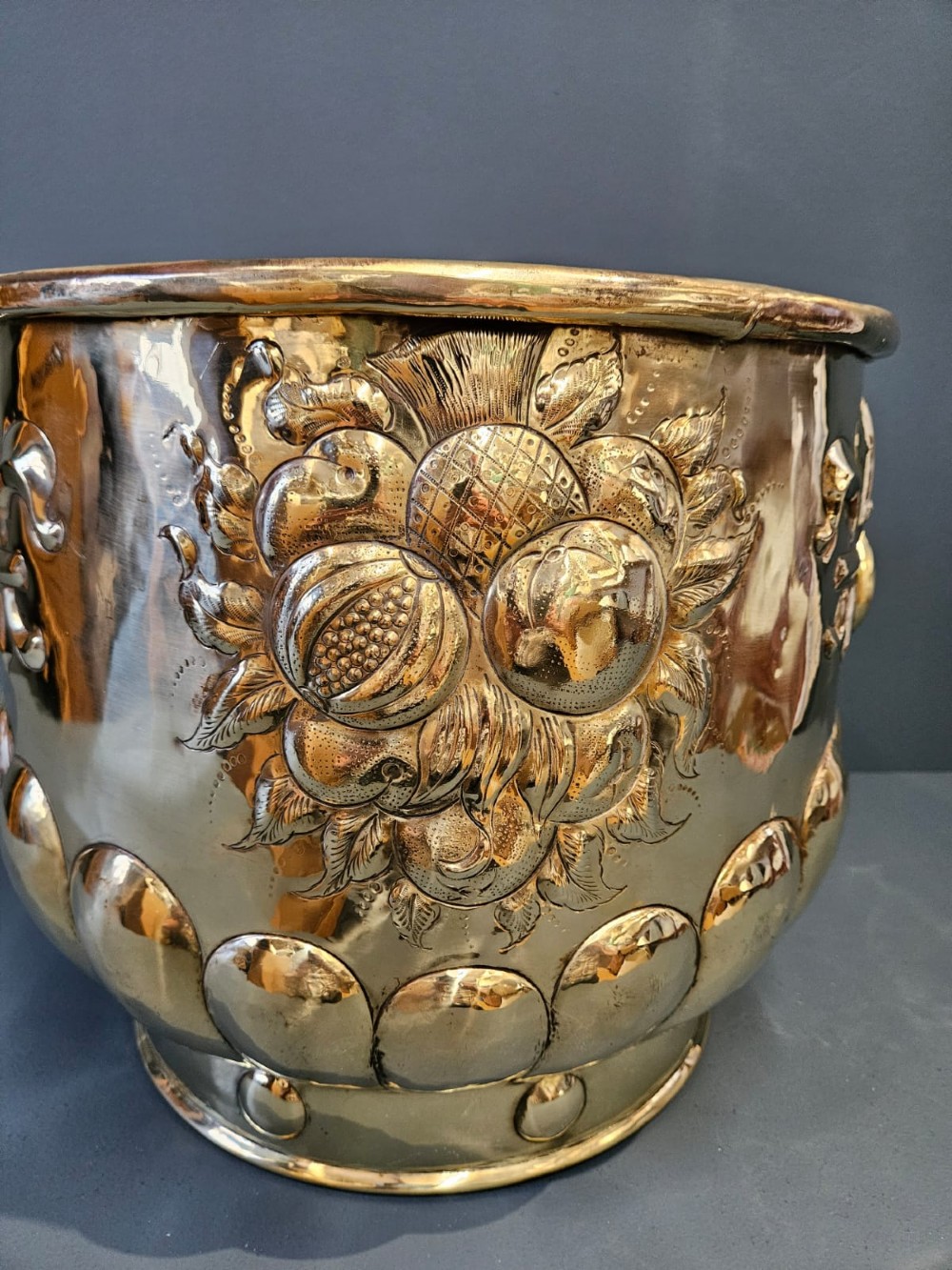 A large polished brass cylindrical planter with an embossed heraldic crest on the front, godrooned base pattern, and side ring handles. View VIII