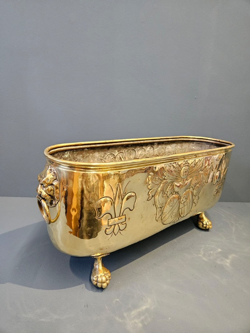 An oblong Victorian brass planter featuring an embossed armorial crest, fleur-de-lis motifs, side ring handles, and cast claw feet. View II