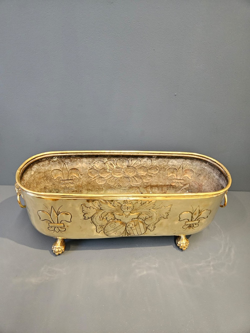 An oblong Victorian brass planter featuring an embossed armorial crest, fleur-de-lis motifs, side ring handles, and cast claw feet. View VI