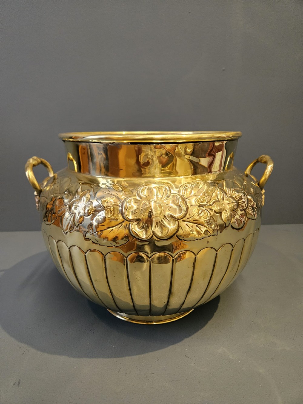 A polished Victorian brass jardiniere planter featuring embossed floral detailing around the upper band, fluted gadrooning on the lower body, and two side handles.