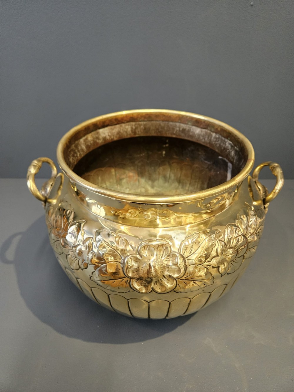 A polished Victorian brass jardiniere planter featuring embossed floral detailing around the upper band, fluted gadrooning on the lower body, and two side handles. View II