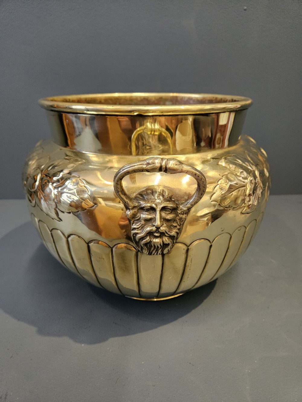 A polished Victorian brass jardiniere planter featuring embossed floral detailing around the upper band, fluted gadrooning on the lower body, and two side handles. View VI