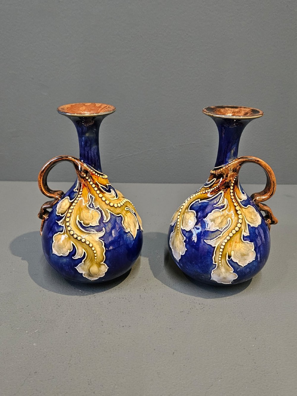 A pair of Victorian Doulton Lambeth stoneware ewers featuring cobalt blue glazed bodies, single handles, and raised foliage decoration in beige and ocher.