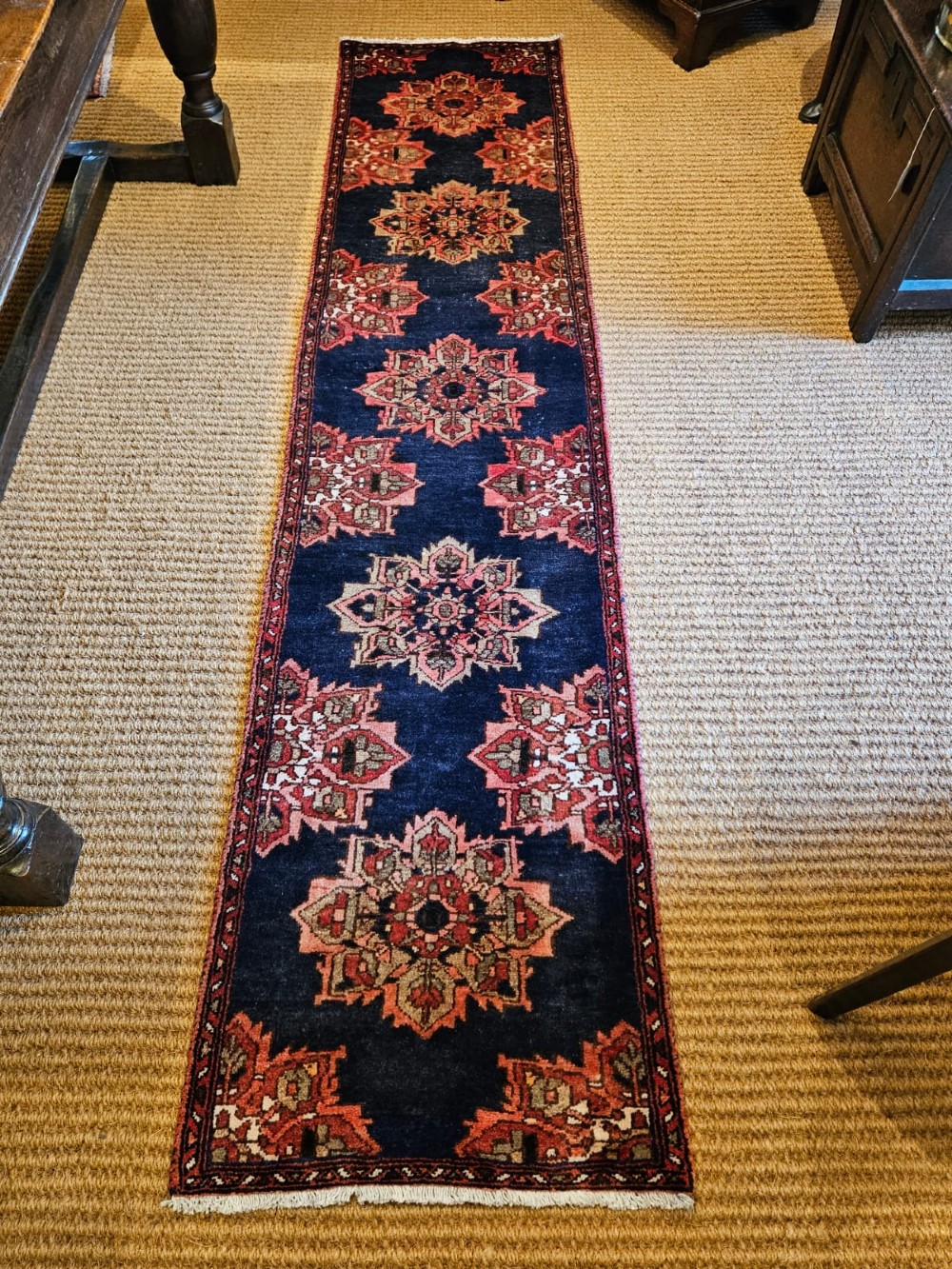 A long narrow navy blue runner rug decorated with a row of repeating geometric star medallions in terracotta and cream tones. View V