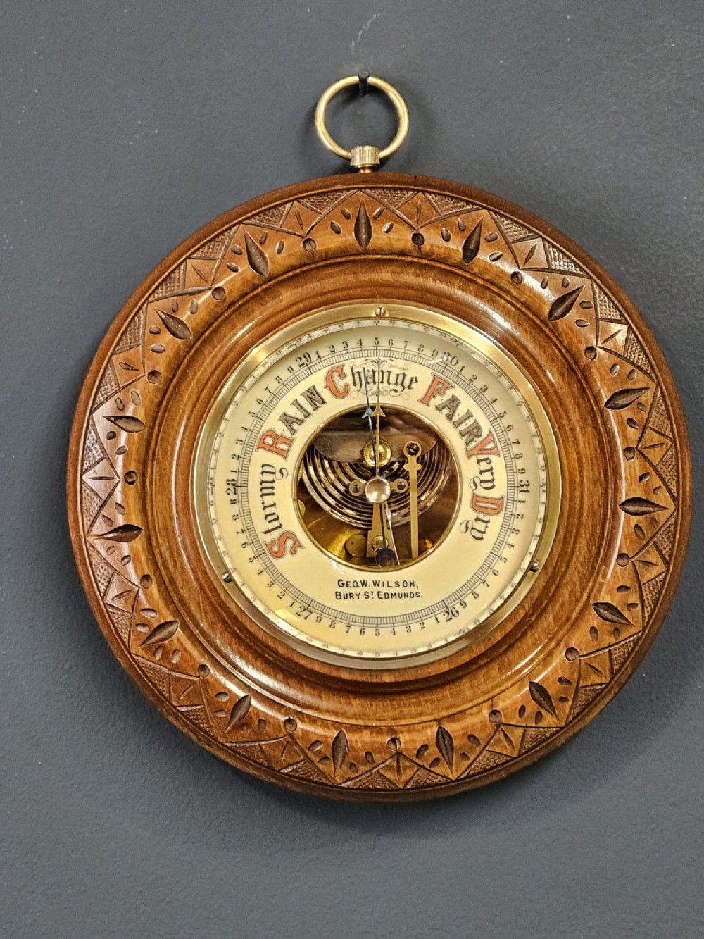 A circular Edwardian aneroid wall barometer featuring a hand-carved walnut frame with geometric repeating patterns, a brass bezel, and a visible central movement surrounded by a cream and red dial.