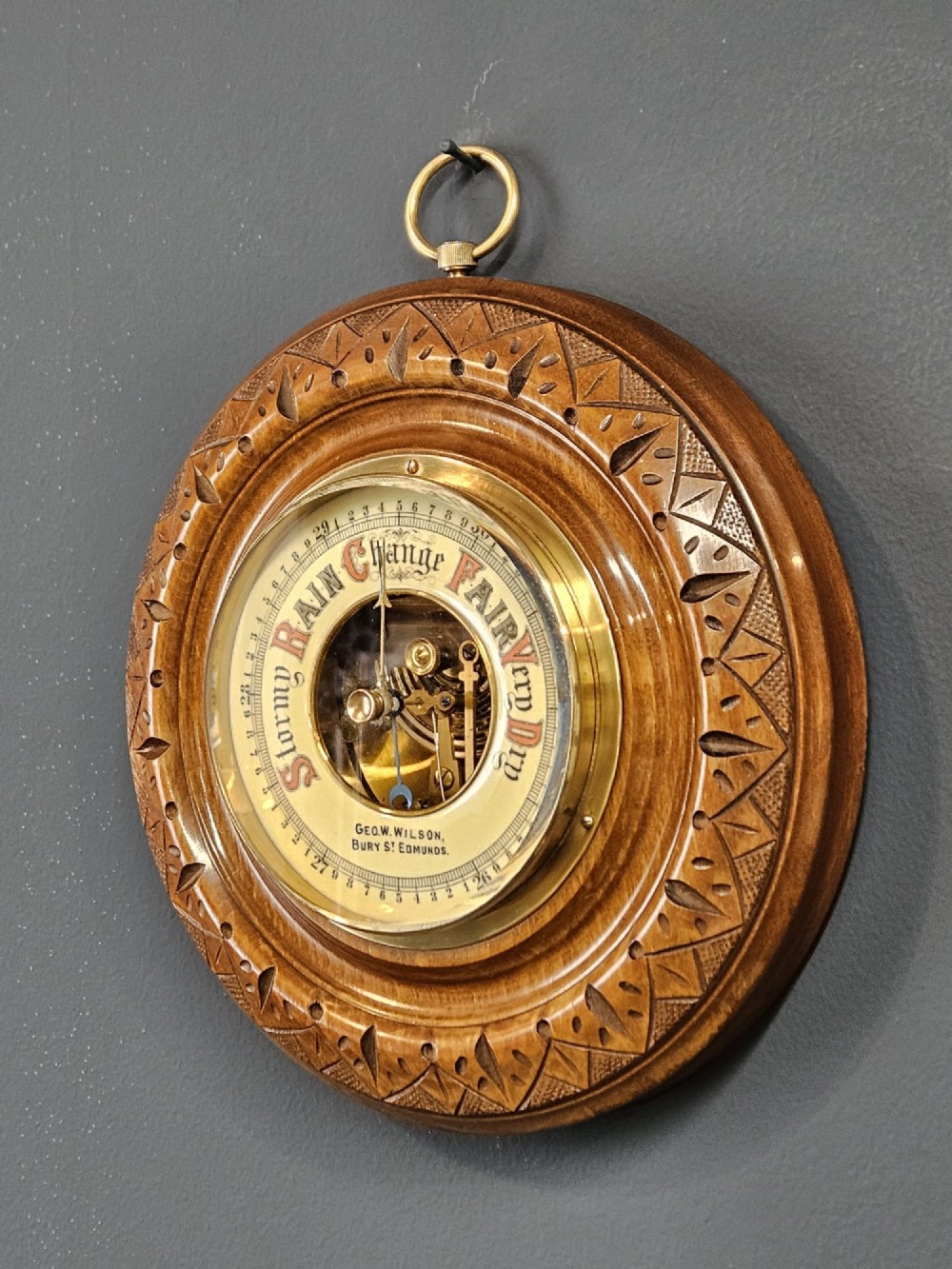 A circular Edwardian aneroid wall barometer featuring a hand-carved walnut frame with geometric repeating patterns, a brass bezel, and a visible central movement surrounded by a cream and red dial. View II