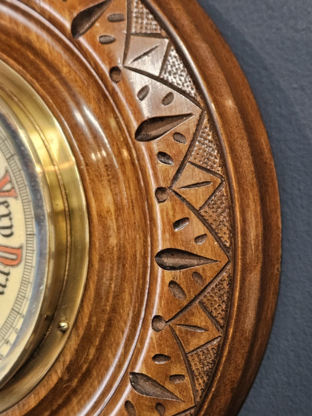 A circular Edwardian aneroid wall barometer featuring a hand-carved walnut frame with geometric repeating patterns, a brass bezel, and a visible central movement surrounded by a cream and red dial. View III