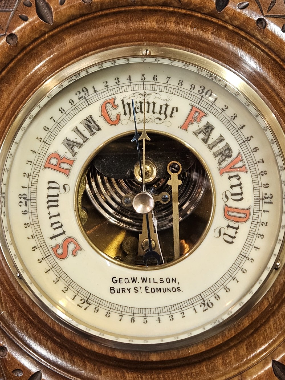 A circular Edwardian aneroid wall barometer featuring a hand-carved walnut frame with geometric repeating patterns, a brass bezel, and a visible central movement surrounded by a cream and red dial. View IV