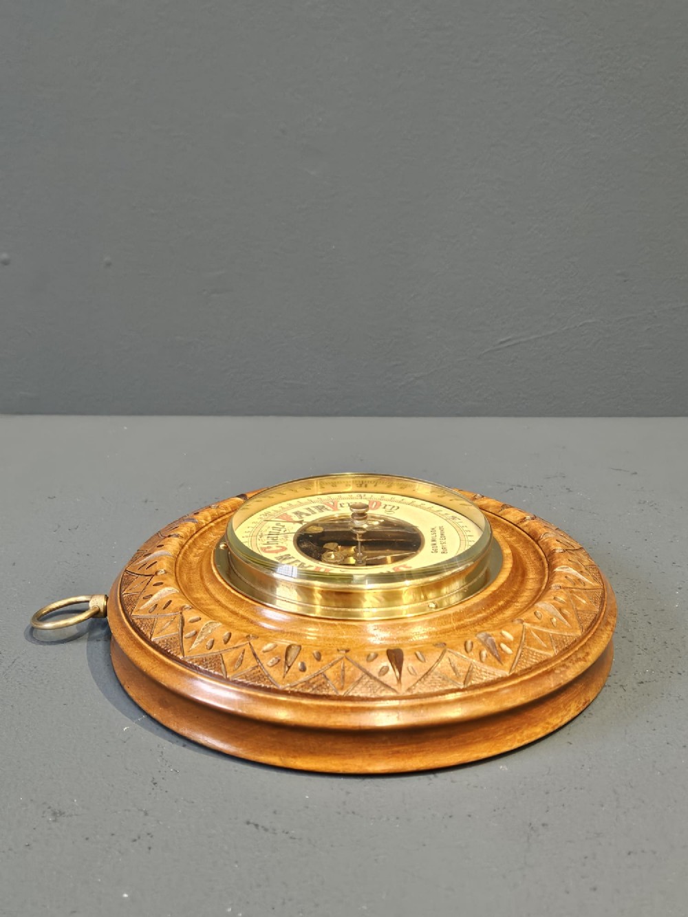 A circular Edwardian aneroid wall barometer featuring a hand-carved walnut frame with geometric repeating patterns, a brass bezel, and a visible central movement surrounded by a cream and red dial. View V