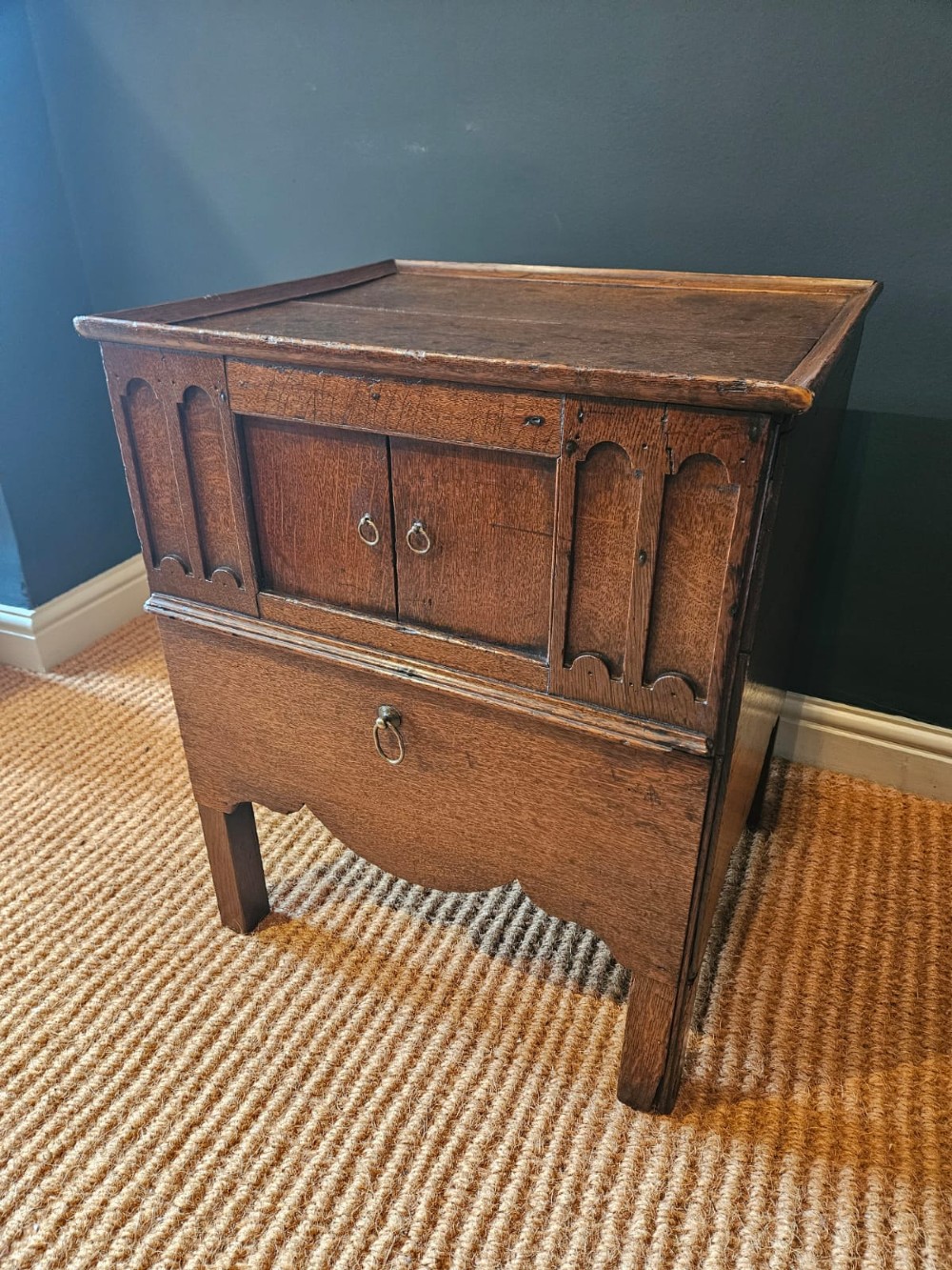 A George III dark oak nightstand featuring a tray top gallery, central cupboard doors flanked by carved arch panels, a lower drawer, and brass ring handles.