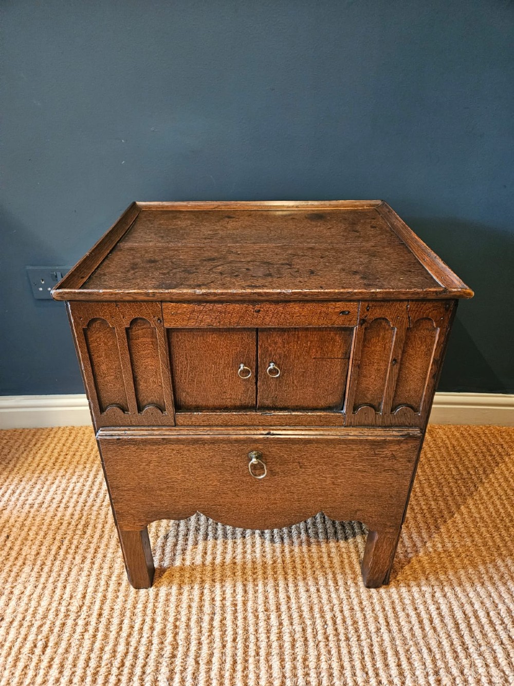 A George III dark oak nightstand featuring a tray top gallery, central cupboard doors flanked by carved arch panels, a lower drawer, and brass ring handles. View II