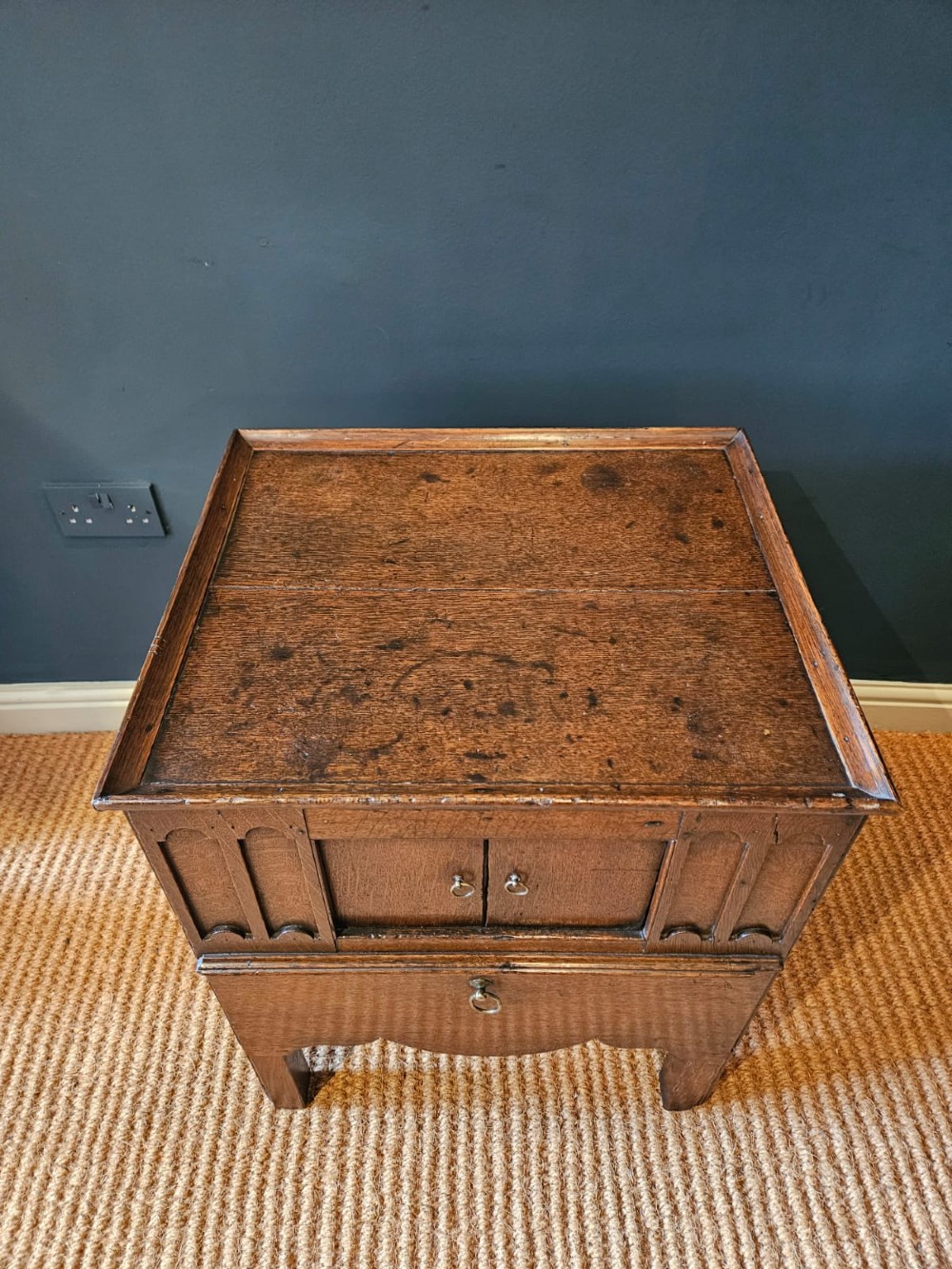 A George III dark oak nightstand featuring a tray top gallery, central cupboard doors flanked by carved arch panels, a lower drawer, and brass ring handles. View III