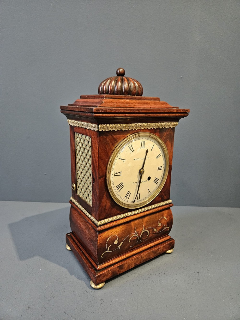 A small Regency period mahogany bracket clock featuring brass inlay, side grilles, a circular dial with Roman numerals, and a carved fluted finial.