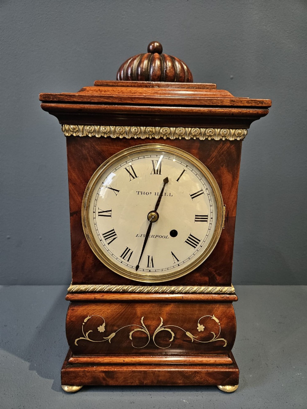 A small Regency period mahogany bracket clock featuring brass inlay, side grilles, a circular dial with Roman numerals, and a carved fluted finial. View II