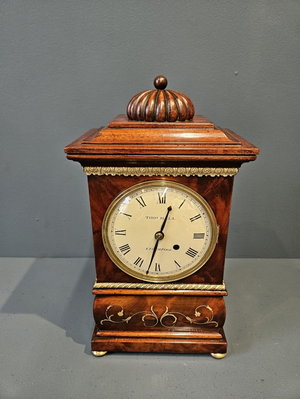 A small Regency period mahogany bracket clock featuring brass inlay, side grilles, a circular dial with Roman numerals, and a carved fluted finial. View III