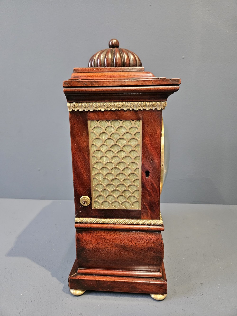 A small Regency period mahogany bracket clock featuring brass inlay, side grilles, a circular dial with Roman numerals, and a carved fluted finial. View IV