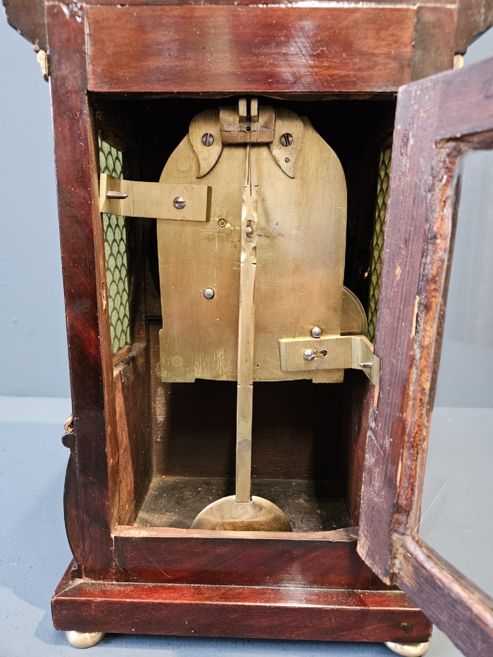 A small Regency period mahogany bracket clock featuring brass inlay, side grilles, a circular dial with Roman numerals, and a carved fluted finial. View V