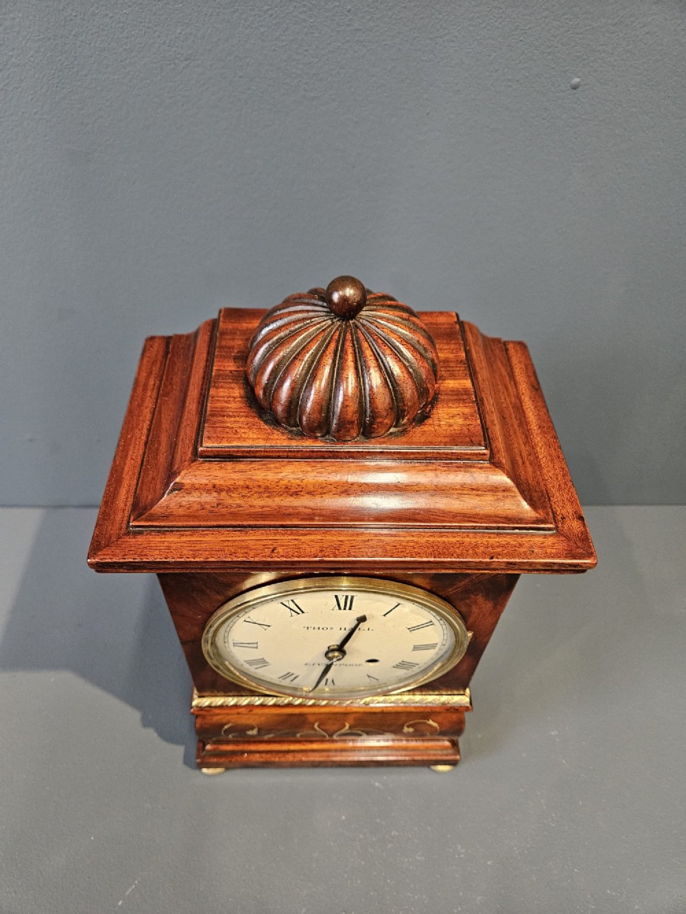 A small Regency period mahogany bracket clock featuring brass inlay, side grilles, a circular dial with Roman numerals, and a carved fluted finial. View VI