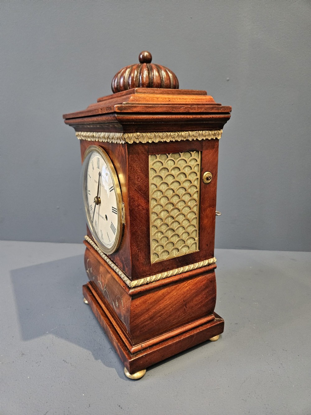 A small Regency period mahogany bracket clock featuring brass inlay, side grilles, a circular dial with Roman numerals, and a carved fluted finial. View IX