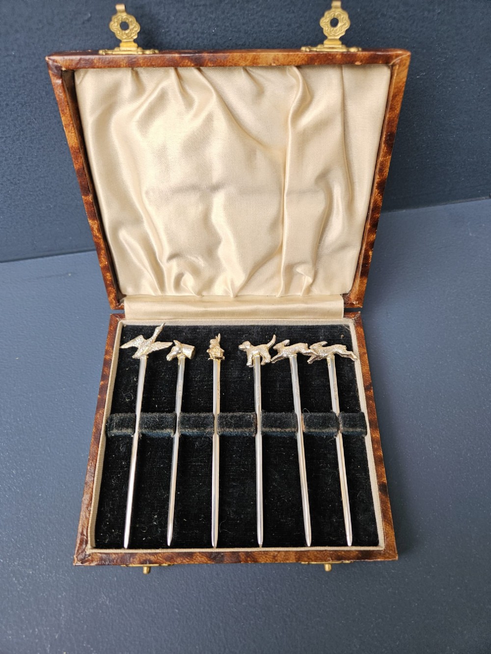 A set of six silver cocktail sticks featuring cast animal finials, arranged inside a fitted presentation box with cream silk and dark velvet lining.
