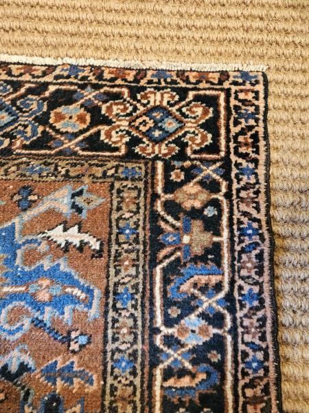 A long hand-knotted wool runner rug featuring central geometric medallion motifs in terracotta brown, indigo blue, and cream with a decorated navy border. View III