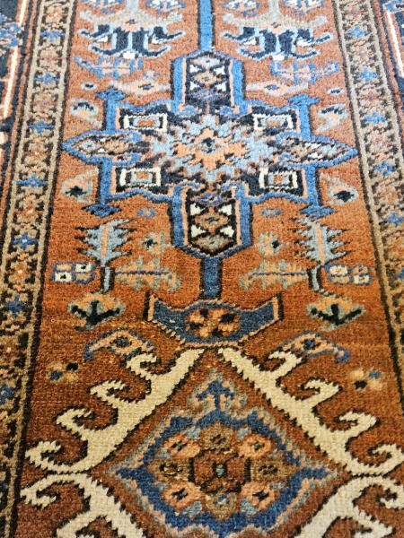 A long hand-knotted wool runner rug featuring central geometric medallion motifs in terracotta brown, indigo blue, and cream with a decorated navy border. View V