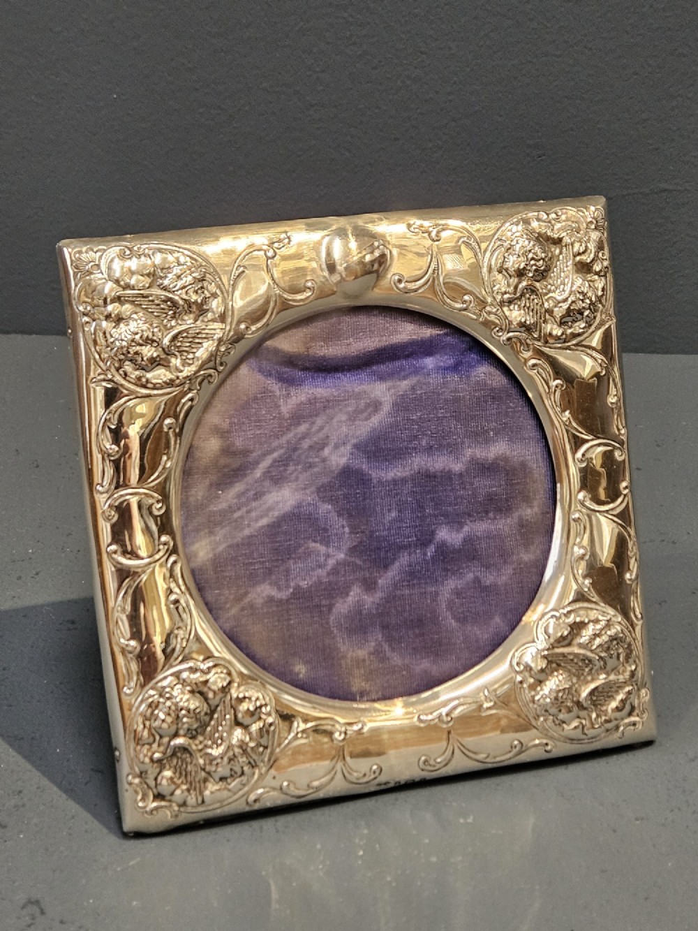 An Edwardian sterling silver square photo frame featuring an oval aperture, embossed cherub heads at each corner, and scrollwork detailing.