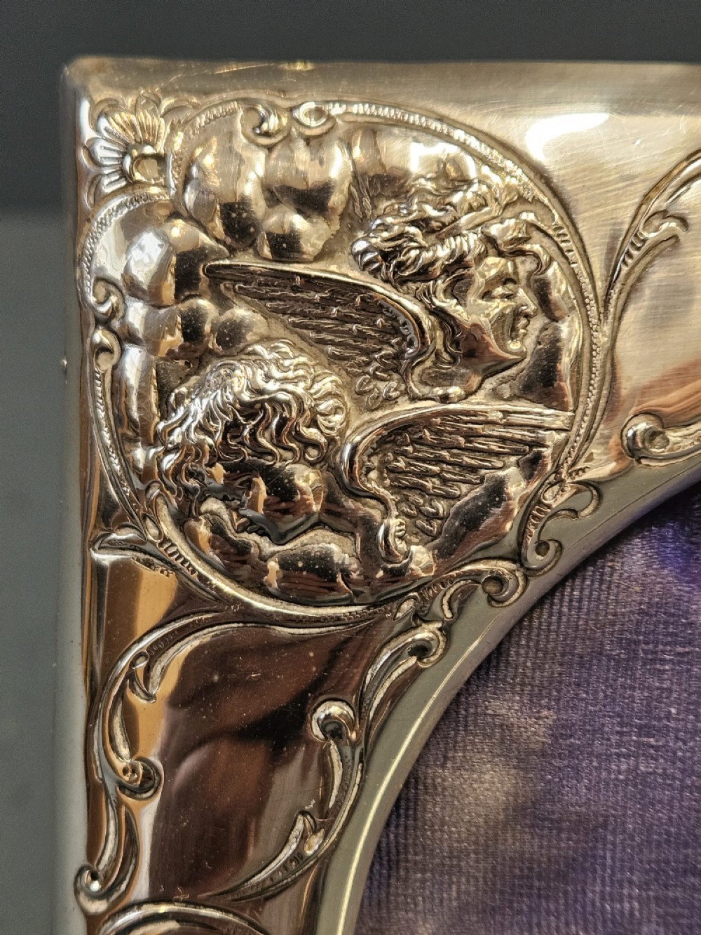 An Edwardian sterling silver square photo frame featuring an oval aperture, embossed cherub heads at each corner, and scrollwork detailing. View II