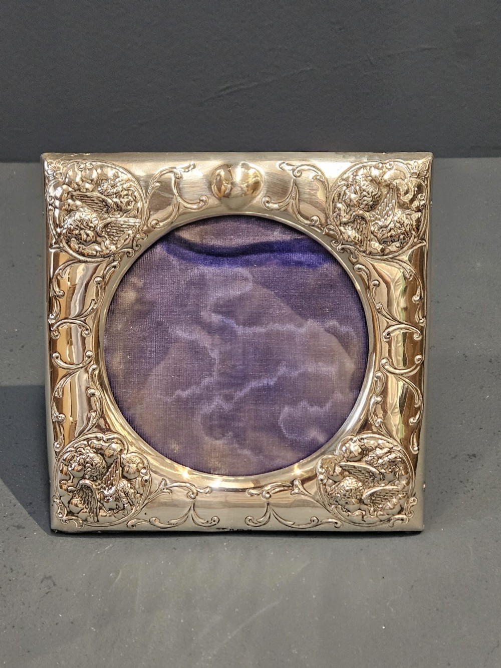 An Edwardian sterling silver square photo frame featuring an oval aperture, embossed cherub heads at each corner, and scrollwork detailing. View III