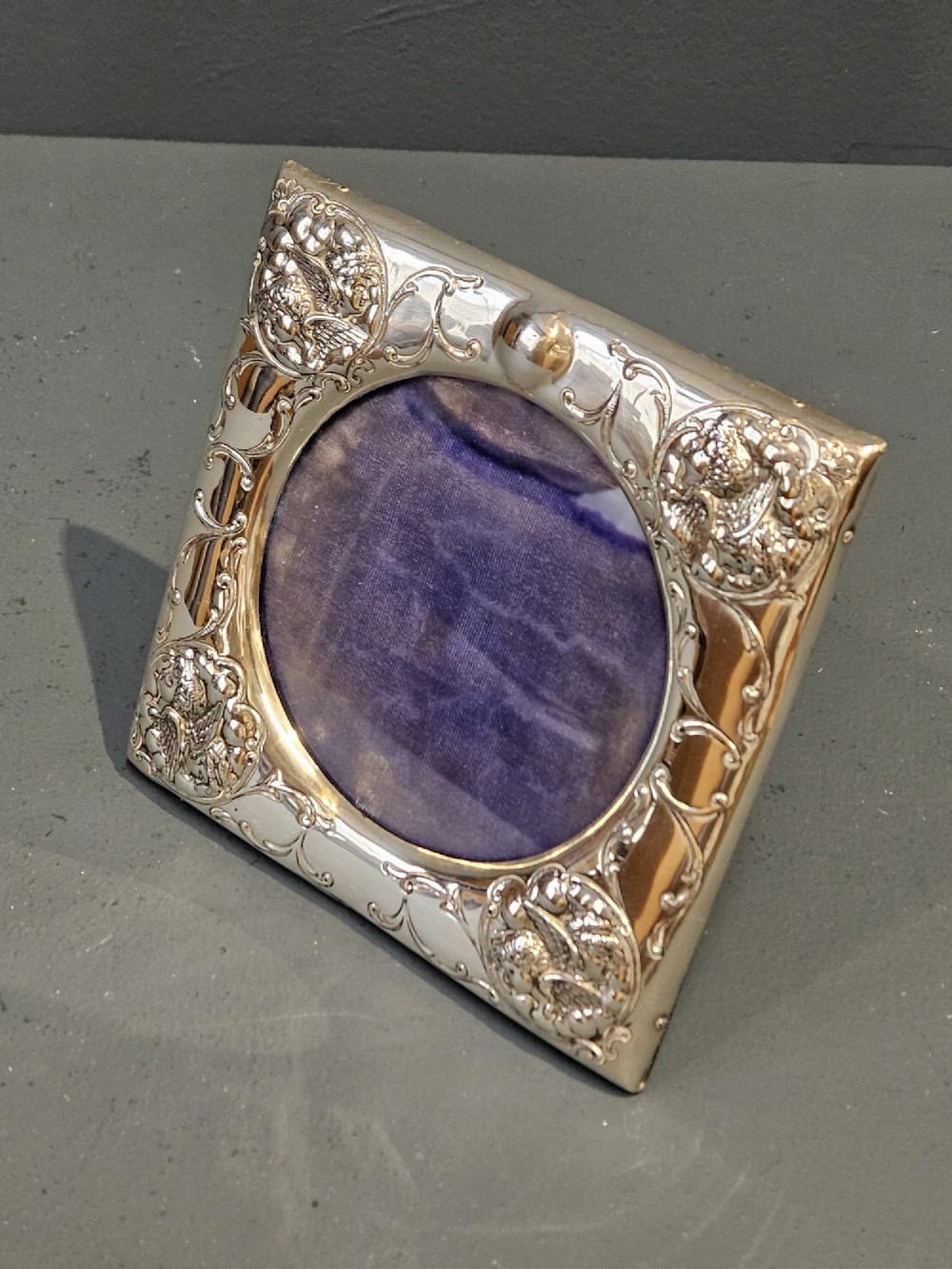 An Edwardian sterling silver square photo frame featuring an oval aperture, embossed cherub heads at each corner, and scrollwork detailing. View VI