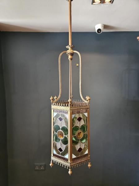 A vertical hexagonal brass hanging lantern featuring stained leaded glass panels with green, purple, and amber floral motifs.