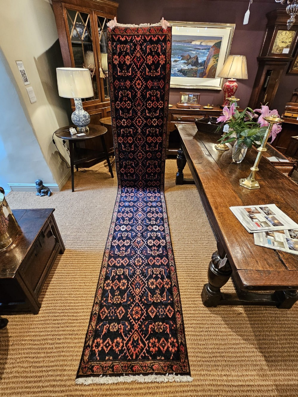 A long hand-knotted wool runner rug featuring repeating geometric diamond patterns in red, cream, and terracotta on a dark navy background with fringe edges.