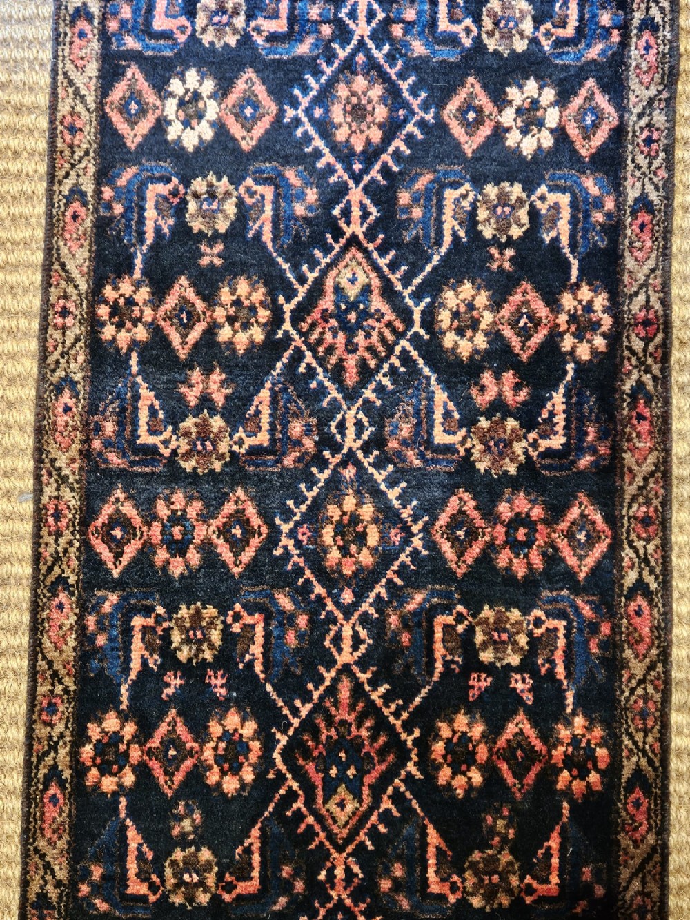 A long hand-knotted wool runner rug featuring repeating geometric diamond patterns in red, cream, and terracotta on a dark navy background with fringe edges. View II