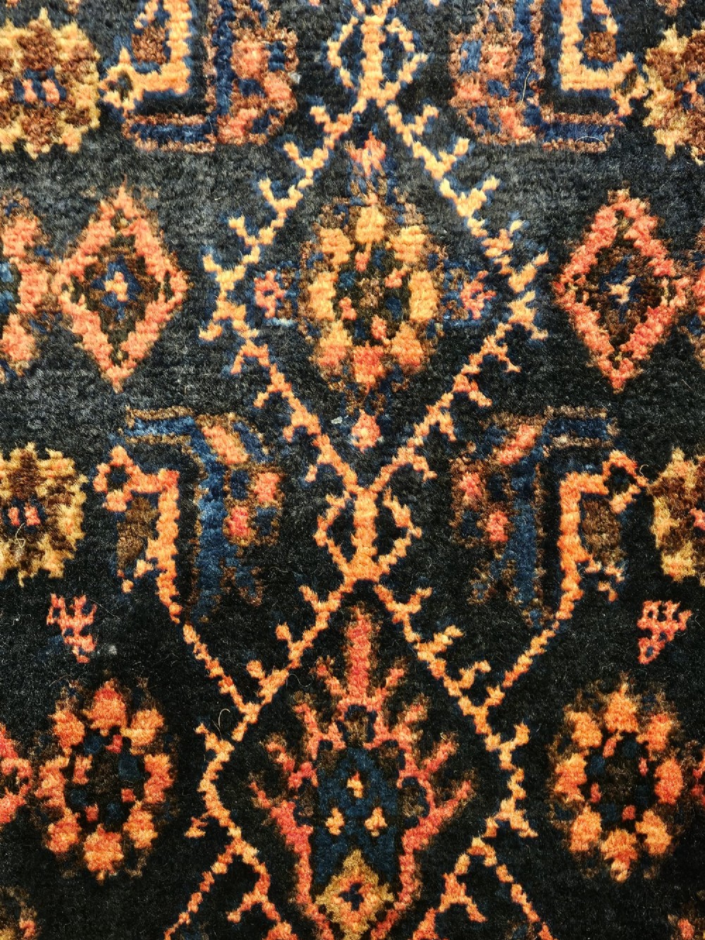 A long hand-knotted wool runner rug featuring repeating geometric diamond patterns in red, cream, and terracotta on a dark navy background with fringe edges. View III