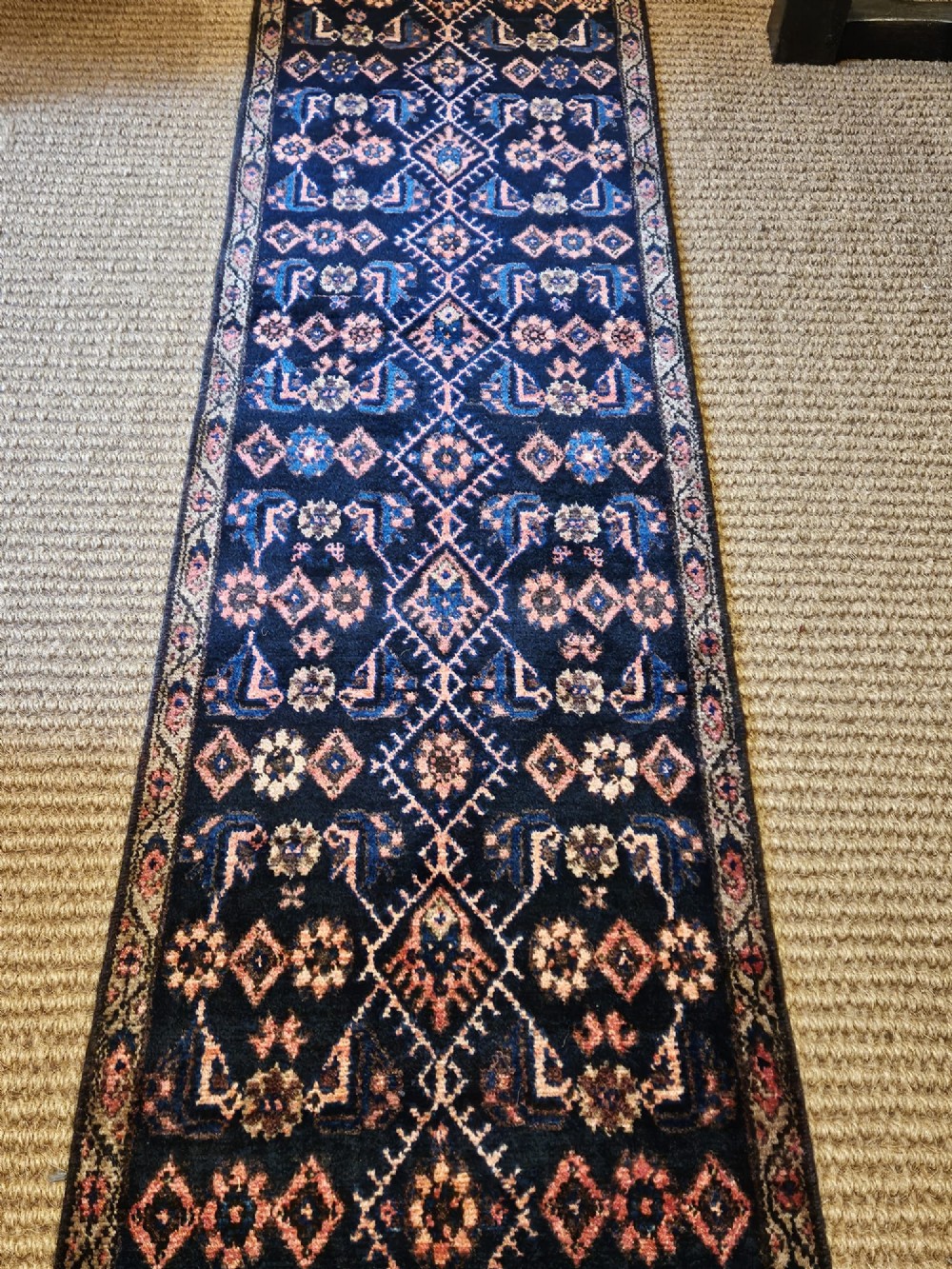 A long hand-knotted wool runner rug featuring repeating geometric diamond patterns in red, cream, and terracotta on a dark navy background with fringe edges. View IV