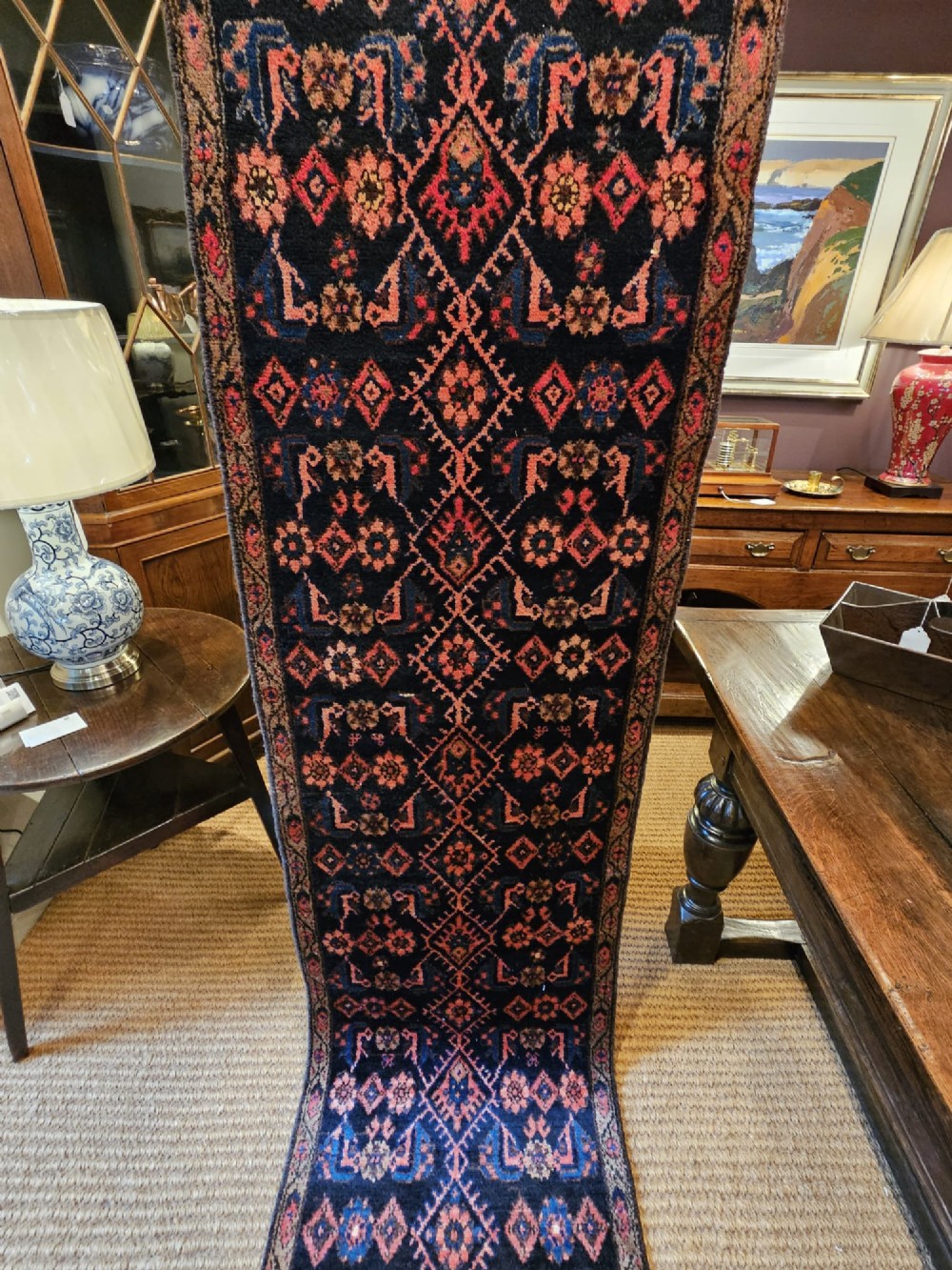 A long hand-knotted wool runner rug featuring repeating geometric diamond patterns in red, cream, and terracotta on a dark navy background with fringe edges. View VI