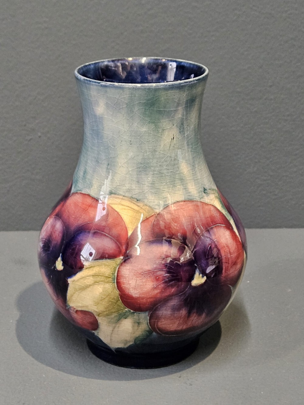 A Moorcroft earthenware vase featuring a bulbous body and flared neck decorated with hand-painted tubelined purple and wine-red pansies beneath a pale blue glaze.
